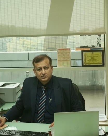 PANKAJ MANCHANDA seated at his executive office desk in a navy suit, a laptop and documents on the desk, glass-walled office behind him