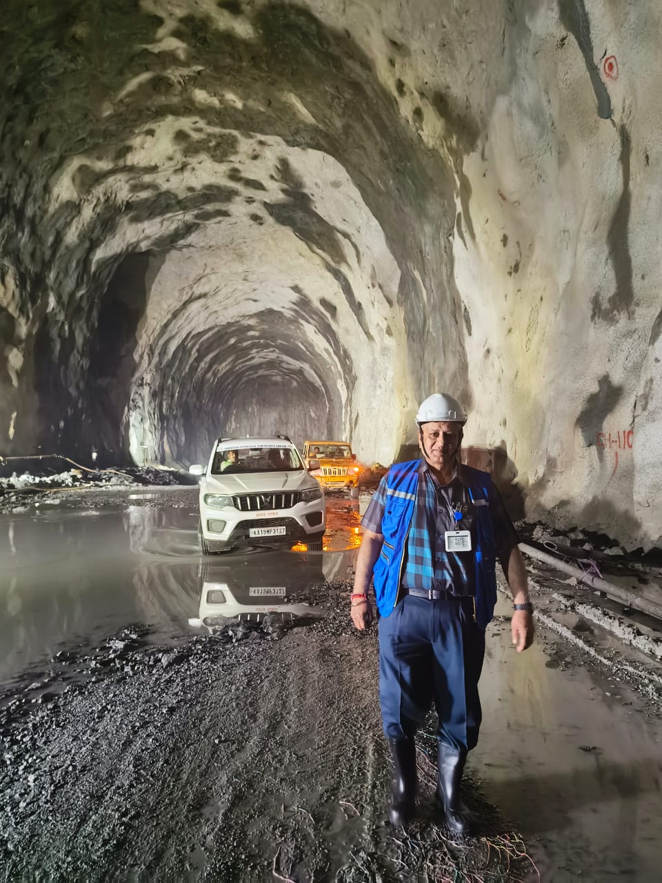PANKAJ MANCHANDA in a white safety helmet and high-visibility vest standing inside a large underground project cavern with vehicles behind him